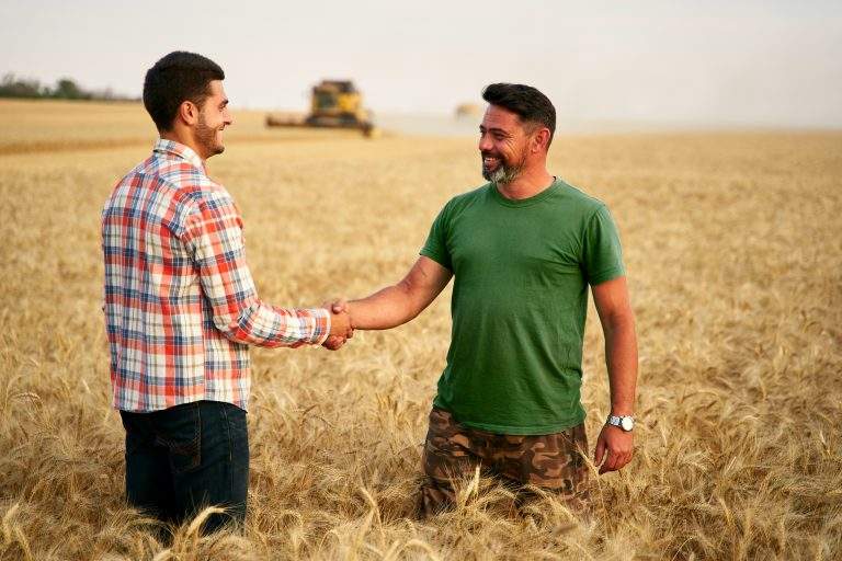 Farmer and agronomist shaking hands in a wheat field after agreement. Agriculture business contract concept. Corporate farmer and landlord rancher negotiate with handshake. Combine harvesters harvest.